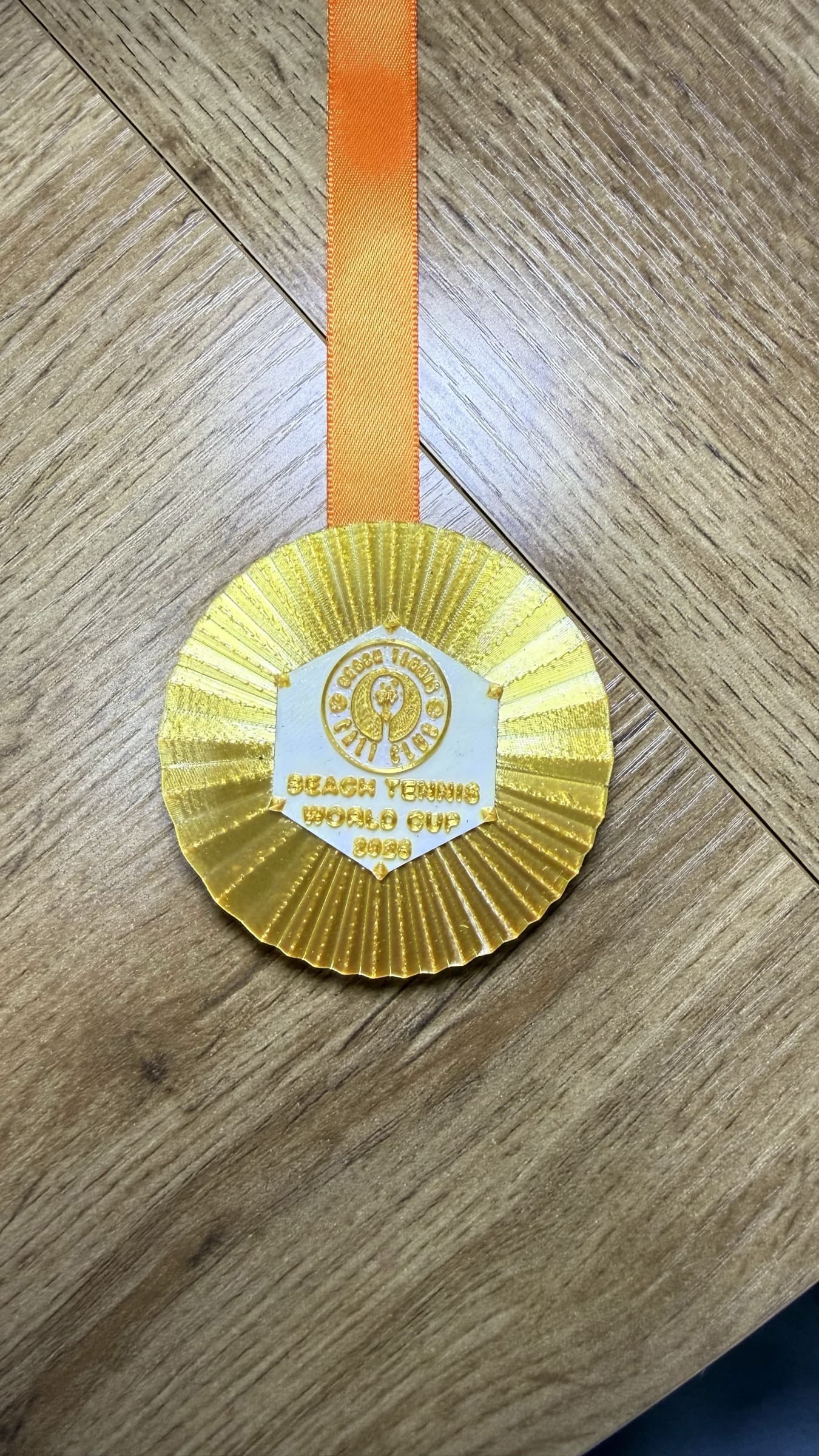 Finished gold Beach Tennis World Cup medal with orange ribbon on a wood surface